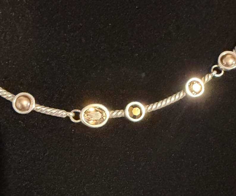 May include: Silver-toned necklace with a twisted rope chain. The necklace features a series of bezel-set, oval and round gemstones in shades of gold and amber. The gemstones are evenly spaced along the chain.