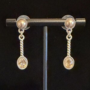 May include: A pair of silver-toned earrings with a gold-colored stud and a dangling spoon-shaped charm. The charm has a twisted stem and an oval-shaped, light-colored stone. The earrings are displayed on a black stand.