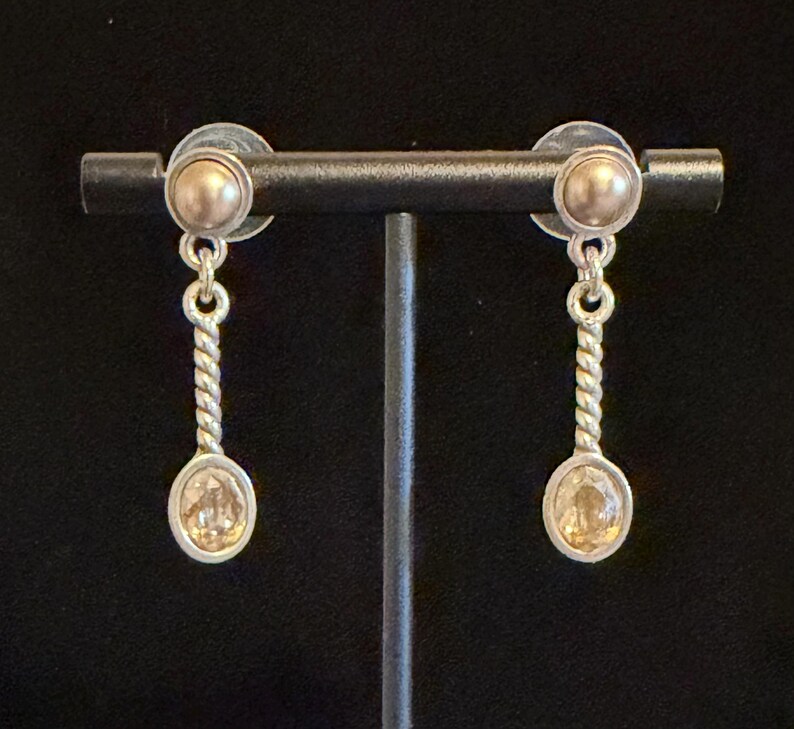 May include: A pair of silver-toned earrings with a gold-colored stud and a dangling spoon-shaped charm. The charm has a twisted stem and an oval-shaped, light-colored stone. The earrings are displayed on a black stand.