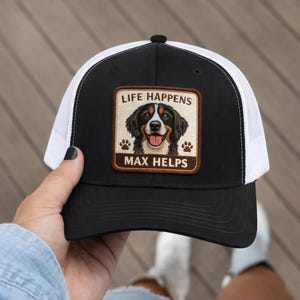 Pode incluir: Boné de caminhoneiro preto e branco com parte traseira em tela e aba curva. A frente apresenta um patch com uma ilustração de cachorro e o texto "LIFE HAPPENS MAX HELPS". O boné está sendo segurado por uma pessoa.
