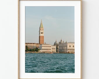 May include: A framed print of Venice, Italy, featuring the iconic St Mark's Campanile and Doge's Palace. The image shows the buildings along the water's edge with a clear blue sky and turquoise water. The print is in a light wood frame.