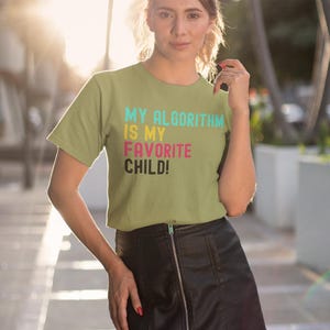 May include: A woman wearing a green t-shirt with the text "MY ALGORITHM IS MY FAVORITE CHILD!" in colorful letters. She is wearing a black leather skirt and has her hair pulled back.