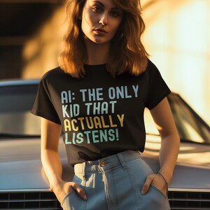 May include: A woman wearing a black t-shirt with the text "AI: THE ONLY KID THAT ACTUALLY LISTENS!" in rainbow colors. She is standing in front of a vintage car and has her hands in her pockets.