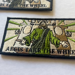 May include: A black and white embroidered patch with a green and white image of a person with long hair and a beard. The person is wearing a green robe and is holding out their hands. The text "AEGIS TAKE THE WHEEL" is embroidered in black and white on the patch.