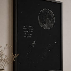 May include: A black framed wall art print featuring a white line drawing of the moon, a small astronaut floating on a tether, and scattered stars. The text reads: I'm an astronaut, you're the moon, I stare at you, I sing to you, I circle you.