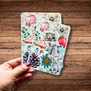 Peut inclure: Deux feuilles d'autocollants avec une variété d'illustrations mignonnes, notamment des théières, des appareils photo, des champignons, une chèvre, une fleur, un cactus, un papillon et une fraise. Les autocollants sont sur un fond à carreaux vert et blanc.