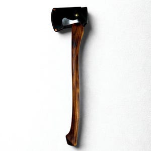 Puede incluir: Hacha con mango de madera oscura y cabeza negra, asegurada por una funda de cuero negro. La cabeza del hacha es plateada. La veta de la madera es visible en el mango. El hacha se muestra sobre un fondo blanco.