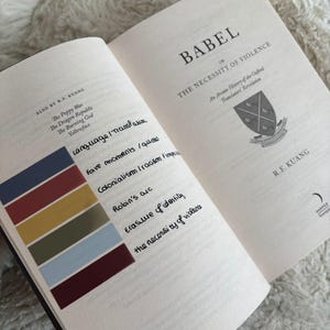 May include: An open book titled "BABEL or The Necessity of Violence" by R.F. Kuang. The left page has handwritten notes and a color-coded sticky note. The right page displays the title and author's name. The book is on a soft, white surface.