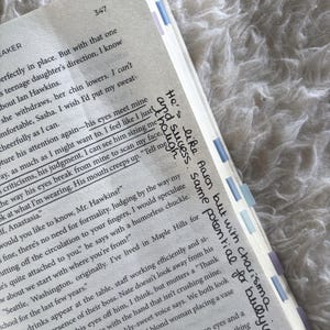 May include: A page from a printed book with black text on white paper, featuring handwritten notes in black ink in the margin. The page includes page number 347 and several light blue and purple adhesive page markers attached to the outer edge.