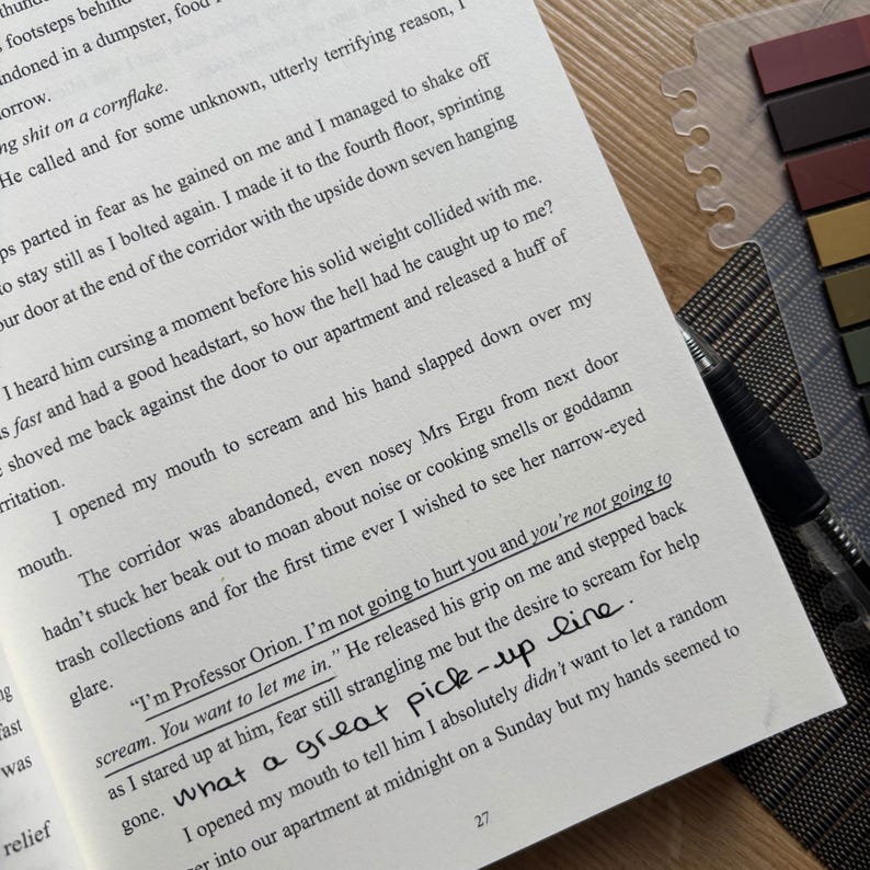 Puede incluir: Libro abierto con texto impreso, un bol&iacute;grafo negro y marcadores de p&aacute;gina coloridos. El texto de la p&aacute;gina incluye las frases "I'm not going to hurt you" y "What a great pick-up line."