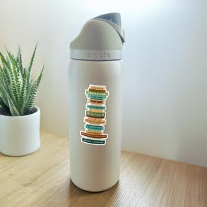 May include: A white Owala water bottle with a gray lid and a stack of colorful stickers. The stickers feature book titles like "Fake Dating" and "Slow Burn". A potted succulent is in the background.