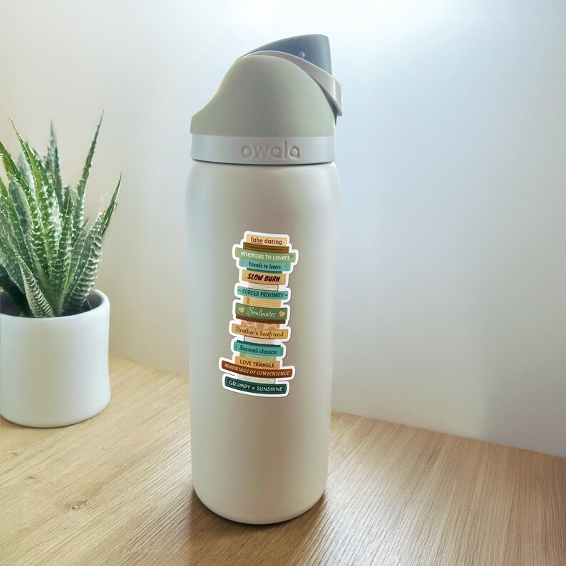 May include: A white Owala water bottle with a gray lid and a stack of colorful stickers. The stickers feature book titles like "Fake Dating" and "Slow Burn". A potted succulent is in the background.
