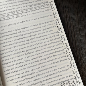 Puede incluir: Libro abierto con texto impreso y notas manuscritas en tinta negra. El texto incluye frases como "Zodiac Academy" y "learning magic". El libro est&aacute; sobre una superficie oscura, con las p&aacute;ginas ligeramente inclinadas.