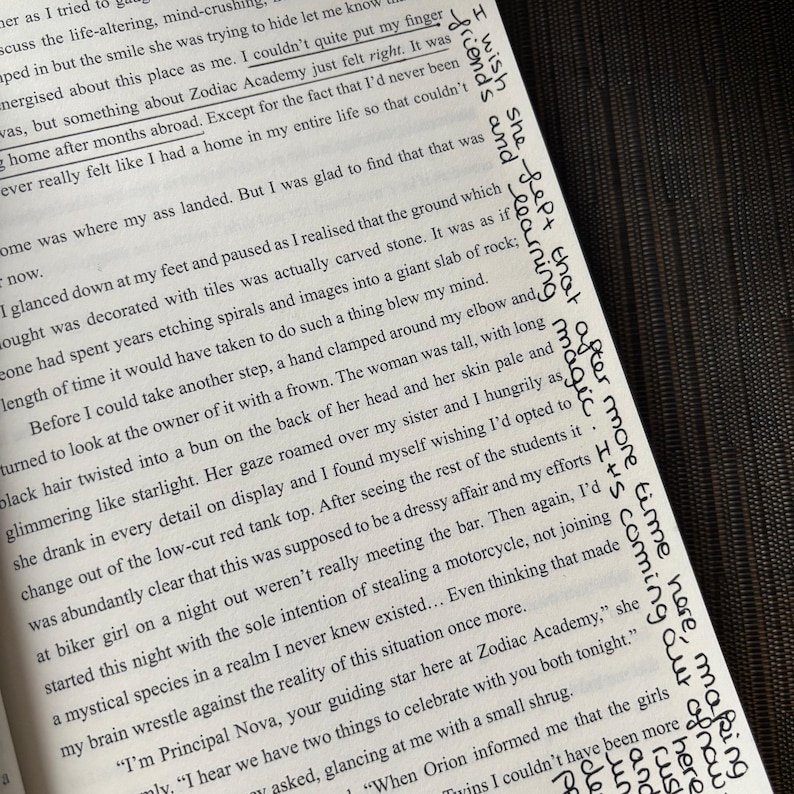 Puede incluir: Libro abierto con texto impreso y notas manuscritas en tinta negra. El texto incluye frases como "Zodiac Academy" y "learning magic". El libro est&aacute; sobre una superficie oscura, con las p&aacute;ginas ligeramente inclinadas.