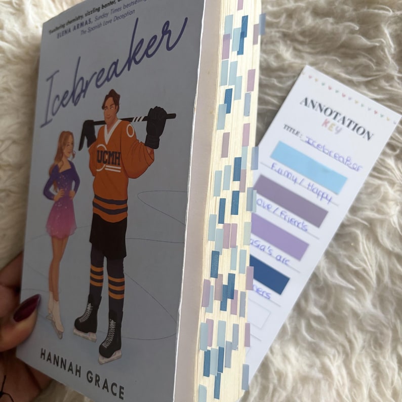 May include: A paperback copy of the book Icebreaker by Hannah Grace with a light blue cover featuring an illustration of a figure skater and a hockey player. The book pages are marked with numerous small blue and purple adhesive page tabs.
