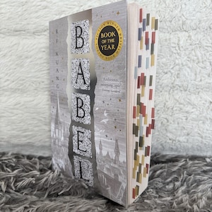 May include: A hardcover book titled "Babel" with a silver and gray cover. The title is in large, ornate letters. The book has a "Book of the Year" sticker and colorful page markers. The author's name is on the spine.