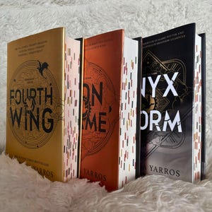 Könnte beinhalten: Drei Hardcover-Bücher mit goldenen, orangefarbenen und schwarzen Einbänden werden gezeigt. Das erste Buch trägt den Titel "Fourth Wing" mit einem goldenen Einband. Das zweite Buch trägt den Titel "Iron Flame" mit einem orangefarbenen Einband. Das dritte Buch trägt den Titel "Nyx Storm" mit einem schwarzen Einband.