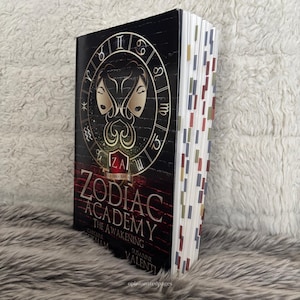 Puede incluir: Un libro de tapa dura titulado "Zodiac Academy: The Awakening" con una cubierta negra que presenta un s&iacute;mbolo del zodiaco dorado y las iniciales "ZA". El libro tiene marcadores de p&aacute;gina coloridos y est&aacute; sobre un fondo suave y texturizado.