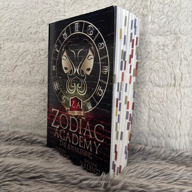 Puede incluir: Un libro de tapa dura titulado "Zodiac Academy: The Awakening" con una cubierta negra que presenta un s&iacute;mbolo del zodiaco dorado y las iniciales "ZA". El libro tiene marcadores de p&aacute;gina coloridos y est&aacute; sobre un fondo suave y texturizado.