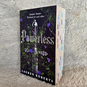 May include: A book titled "Powerless" by Lauren Roberts. The cover shows a silver sword design with purple flowers and green foliage against a dark background. The book's pages are marked with colourful sticky notes.