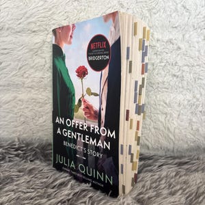 May include: A paperback book titled "An Offer From A Gentleman" by Julia Quinn. The cover features a romantic scene with a red rose and the Netflix Bridgerton logo. The book's pages are marked with colorful tabs.