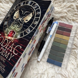 Puede incluir: Un libro de tapa dura titulado "Zodiac Academy: Awakening" con una cubierta negra y dorada con s&iacute;mbolos del zodiaco. El libro tiene pesta&ntilde;as de p&aacute;gina coloridas, un bol&iacute;grafo negro, un bol&iacute;grafo blanco, un juego de muestras de color y una regla transparente.