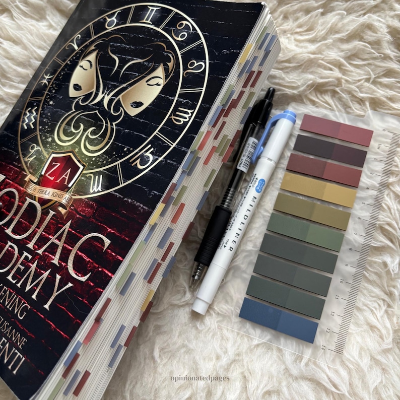 Puede incluir: Un libro de tapa dura titulado "Zodiac Academy: Awakening" con una cubierta negra y dorada con s&iacute;mbolos del zodiaco. El libro tiene pesta&ntilde;as de p&aacute;gina coloridas, un bol&iacute;grafo negro, un bol&iacute;grafo blanco, un juego de muestras de color y una regla transparente.