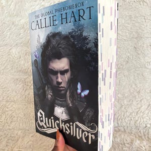 Può includere: Un libro tascabile intitolato "Quicksilver" di Callie Hart. La copertina presenta un uomo con i capelli scuri e una lacrima, con farfalle e una spada. Il dorso del libro ha segnalibri colorati.