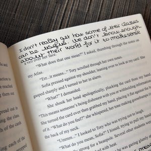 Puede incluir: P&aacute;ginas de un libro abierto con texto impreso y notas manuscritas en tinta negra. El texto incluye frases como "I don't really get how some of their classes can be useful." El libro est&aacute; abierto sobre una superficie texturizada.