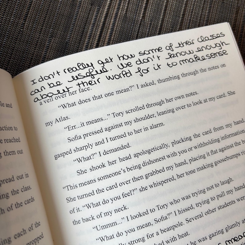Puede incluir: P&aacute;ginas de un libro abierto con texto impreso y notas manuscritas en tinta negra. El texto incluye frases como "I don't really get how some of their classes can be useful." El libro est&aacute; abierto sobre una superficie texturizada.