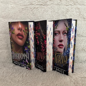 May include: Three hardcover books with illustrated covers. The book spines are white with vertical stripes in various colors. The book covers feature portraits of women and floral designs. The titles are "Belladonna", "Glove", and "Teria".