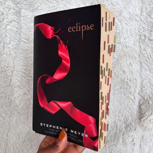 May include: A hardcover book titled "eclipse" by Stephenie Meyer. The cover is black with a flowing red ribbon design. The book's spine has colorful rectangular bookmarks.