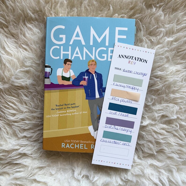 May include: A paperback book titled "Game Change" with a light blue cover and illustrations of two men. Beside it is a bookmark with annotation labels. The book is by Rachel Reid.