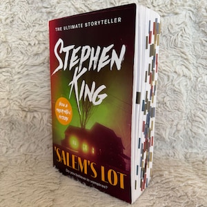 Op de afbeelding: Een hardcover boek getiteld "'Salem's Lot" van Stephen King. De omslag toont een illustratie van een huis in groene en oranje tinten. De titel en de naam van de auteur zijn prominent aanwezig. De tekst "Now a Major Motion Picture" is ook zichtbaar.