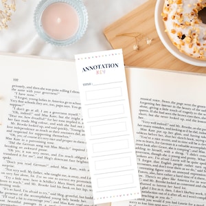 Puede incluir: Un marcapáginas con "ANNOTATION KEY" y espacio para un título, sobre una página de libro. Un donut glaseado y una vela también son visibles, evocando la lectura.