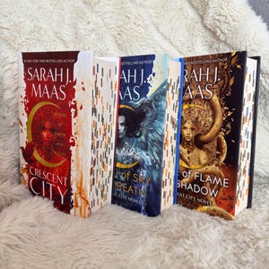 May include: A stack of four hardcover books by Sarah J. Maas. The book covers feature illustrations in shades of red, blue, and gold. Titles include "House of Earth and Blood" and "Of Flame and Shadow". The spines have a patterned design.