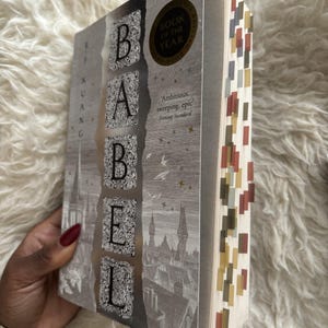 May include: A hardcover book titled "Babel" with a silver cover featuring the title in large, ornate letters. The book has a "Book of the Year" sticker and colorful page markers. The cover also includes the author's name and a quote.