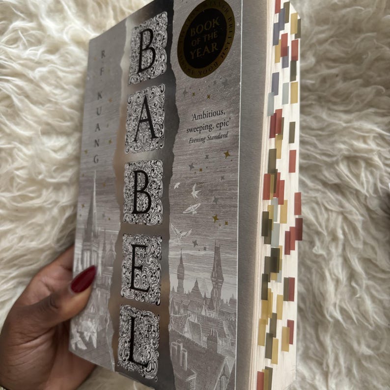 May include: A hardcover book titled "Babel" with a silver cover featuring the title in large, ornate letters. The book has a "Book of the Year" sticker and colorful page markers. The cover also includes the author's name and a quote.