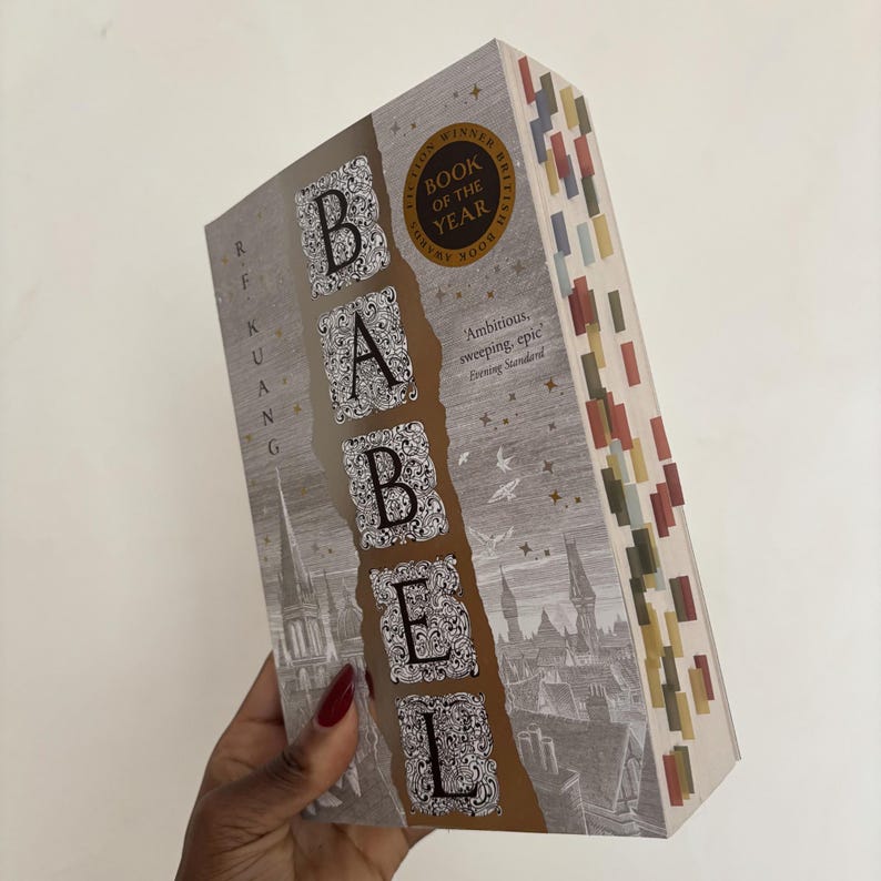 May include: A hardcover book titled "Babel" with the author's name, R.F. Kuang, visible. The cover features the title in ornate lettering, a cityscape illustration, and a "Book of the Year" award seal. The book has colorful page markers.