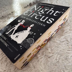 May include: A copy of 'The Night Circus' by Erin Morgenstern, featuring a black cover with white illustrations and the title in white. The book has colourful page markers and is lying on a white, textured surface.
