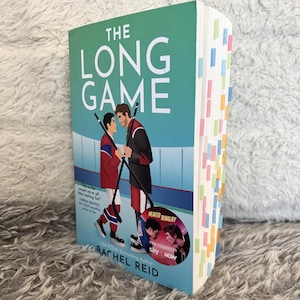 May include: A book titled "The Long Game" by Rachel Reid. The cover shows two hockey players facing each other with hockey sticks. The book has a green background and colourful sticky notes on the side.