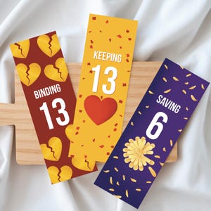 Pode incluir: Três marcadores coloridos com texto e gráficos. Um é bordô com gráficos de coração partido e as palavras "BINDING 13". O segundo é amarelo com um coração vermelho e as palavras "KEEPING 13". O terceiro é roxo com um gráfico de flor e as palavras "SAVING 6".