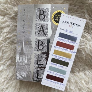 May include: A hardcover book titled "Babel" with a silver cover and ornate lettering. Beside it is a bookmark with the title and annotation key labels like "Language/Translation" and "Robin's arc".