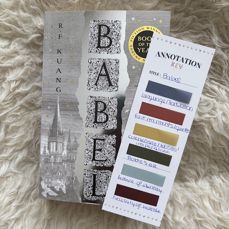 May include: A hardcover book titled "Babel" with a silver cover and ornate lettering. Beside it is a bookmark with the title and annotation key labels like "Language/Translation" and "Robin's arc".