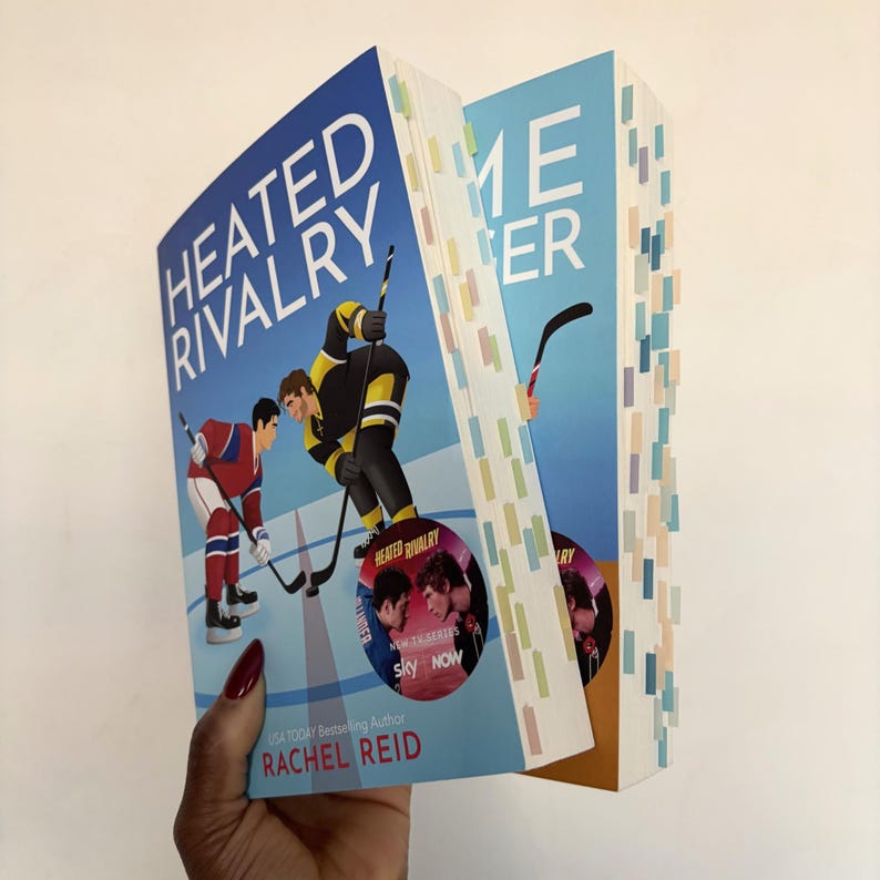 May include: Two books titled "Heated Rivalry" and "Game Changer" are held up. The "Heated Rivalry" book cover features two hockey players facing each other. The book is by Rachel Reid. The second book has a light blue cover with a hockey stick.
