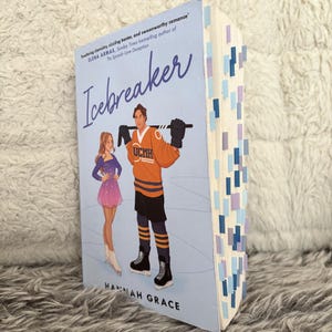May include: A paperback book titled Icebreaker by Hannah Grace with a light blue cover featuring an illustration of a figure skater and a hockey player. The book pages are marked with numerous small blue and purple adhesive page tabs.