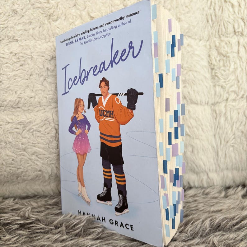 May include: A paperback book titled Icebreaker by Hannah Grace with a light blue cover featuring an illustration of a figure skater and a hockey player. The book pages are marked with numerous small blue and purple adhesive page tabs.