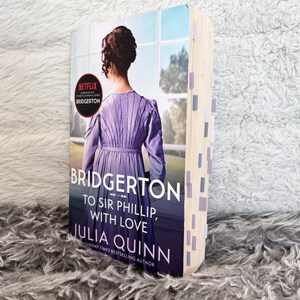 May include: A paperback book titled "Bridgerton: To Sir Phillip, With Love" by Julia Quinn. The cover features a woman in a purple dress, with a Netflix logo. The book's pages have several sticky notes.