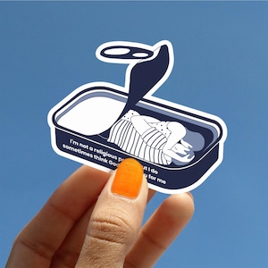 May include: A sticker featuring a sardine can design with a person inside. The can is dark blue with white text that reads "I'm not a religious person but I do sometimes think God is for me."