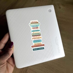 May include: A white Rakuten Kobo e-reader with a stack of colorful stickers. The stickers feature text like "fake dating," "enemies to lovers," and "marriage of convenience." The e-reader is held by a person with a dark red nail polish.